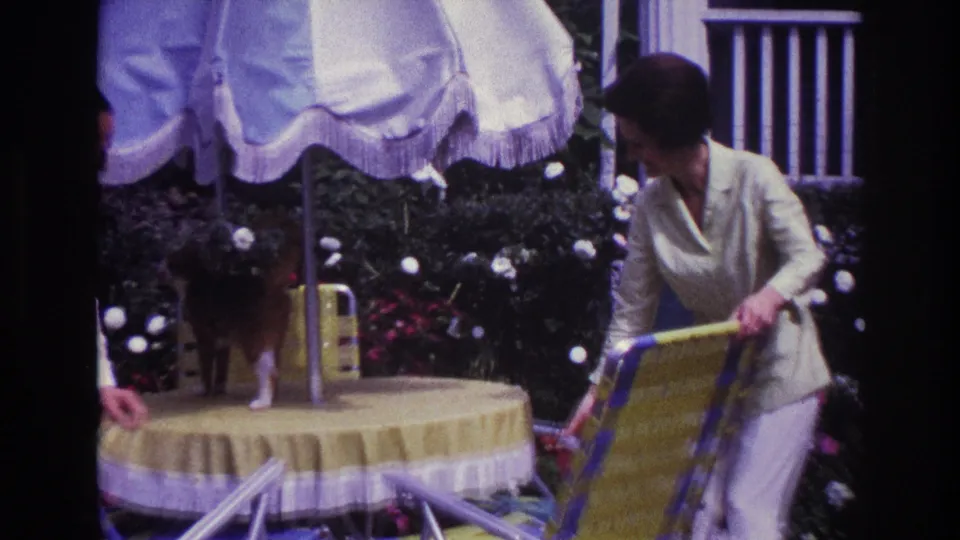 1967 Rockaway Beach home-movie frame of adults arranging a patio table and striped chair under a white umbrella in a garden setting.
