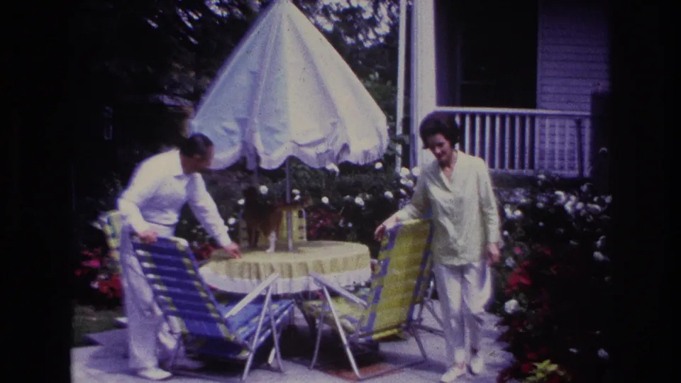 1967 Rockaway Beach home-movie frame of two adults beside a round patio table under a white umbrella, with flowers and a porch in the background.