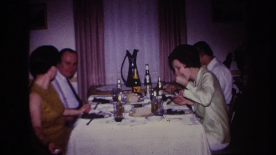 1967 home-movie frame from Rockaway Beach showing adults seated around an indoor dinner table with bottles, plates, food, and curtains behind them.