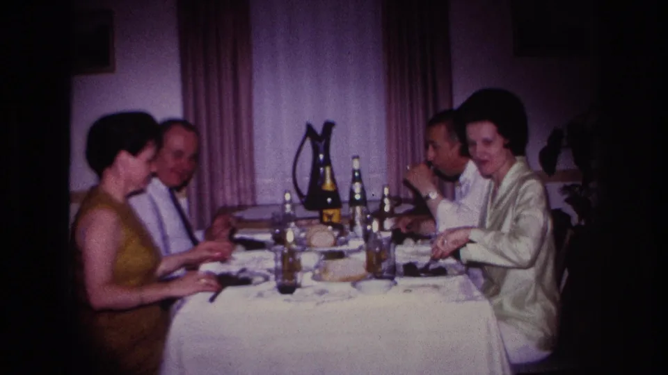1967 Rockaway Beach home interior showing two women and two men eating at a white-covered table with bottles, glasses, and a pitcher.