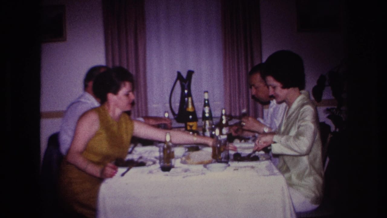 1967 Rockaway Beach home-movie frame showing four dressed adults at an indoor dinner table with bottles, glasses, plates, and curtains behind them.