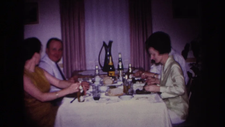 1967 Rockaway Beach home-movie frame showing adults around a dining table, with a man facing the camera and two women partly turned toward the meal.
