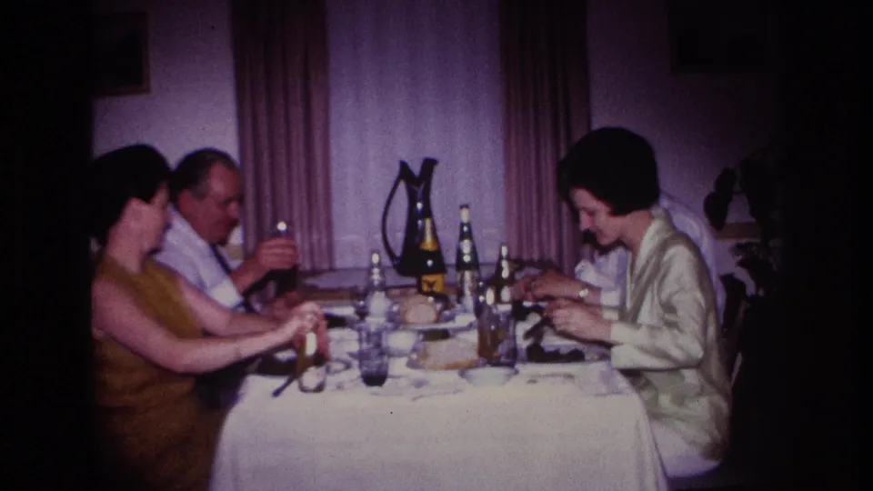 1967 Rockaway Beach home-movie frame of four adults at an indoor meal, with bottles, glasses, a white tablecloth, and purple curtains visible.