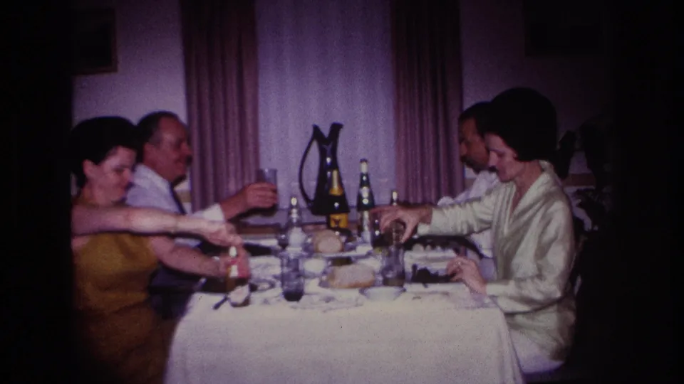 1967 Rockaway Beach home-movie frame showing adults at a meal, including a raised glass during a toast-like moment.