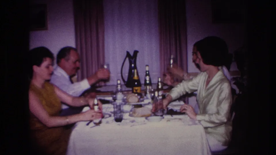 1967 Rockaway Beach home-movie frame with a man lifting a bottle beside seated adults at an indoor dining table.
