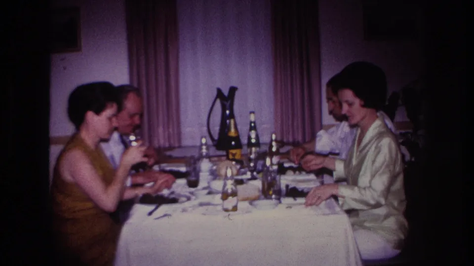1967 home-movie frame from Rockaway Beach showing two couples seated at dinner in a private interior.