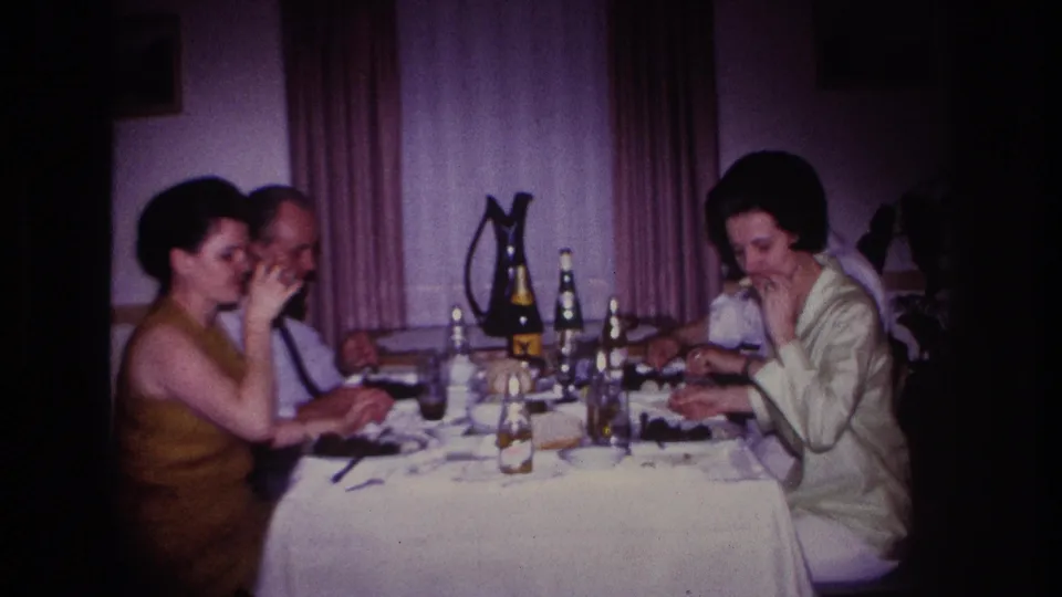 Rockaway Beach 1967 home-movie frame showing adults dining under low interior light with bottles and glasses on the table.