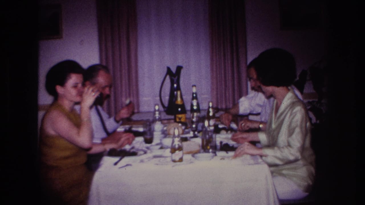 1967 Rockaway Beach home-movie frame of two couples seated at a restaurant table with drinks, plates, and curtains in the background.