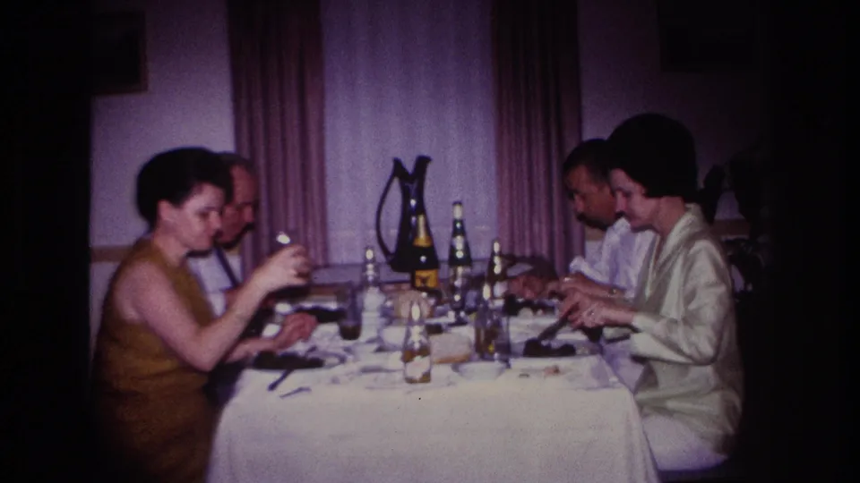 1967 Rockaway Beach home-movie frame of adults dining together, with a woman holding a drink at a white-clothed table.