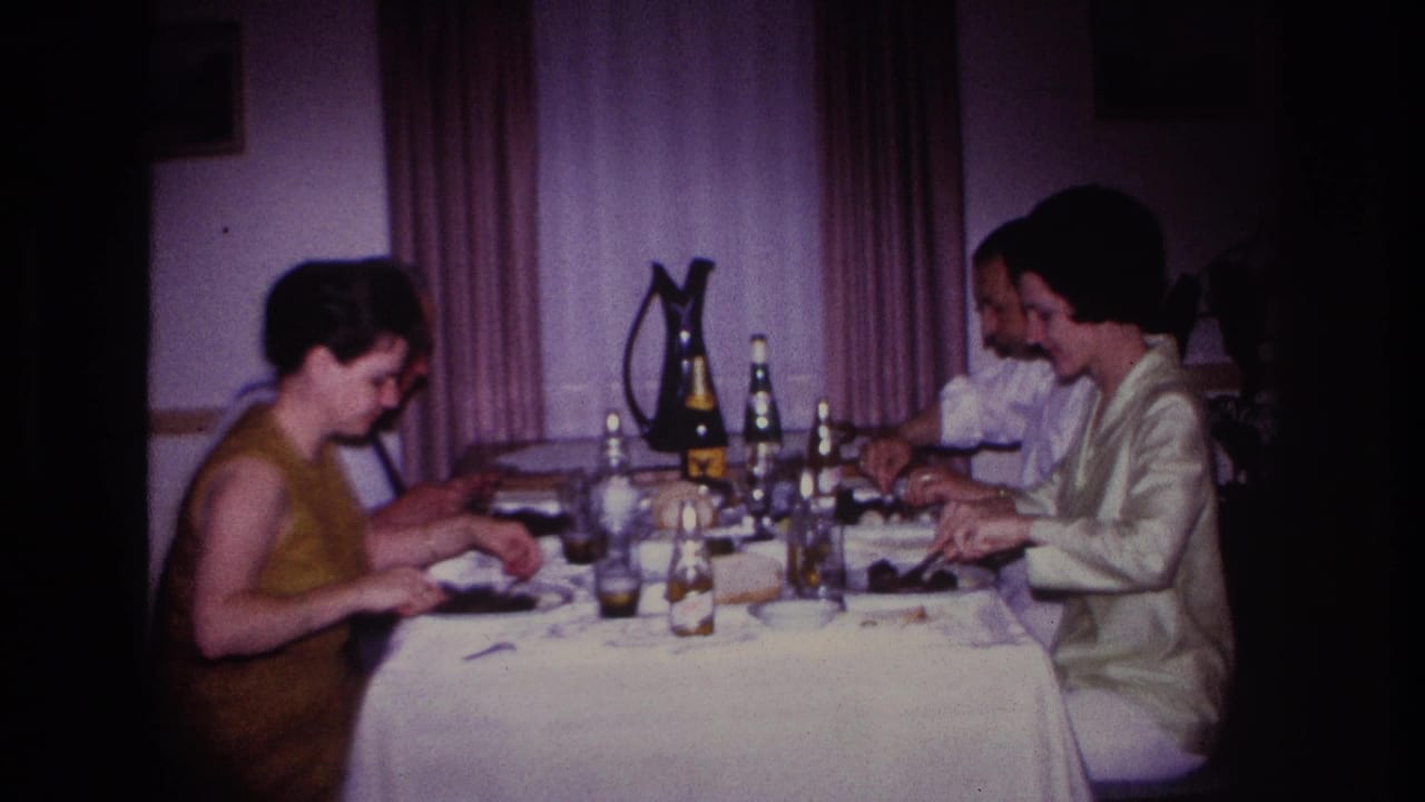 1967 Rockaway Beach home-movie frame showing four adults dining indoors at a formally set table with bottles, glasses, plates, and curtains behind them.