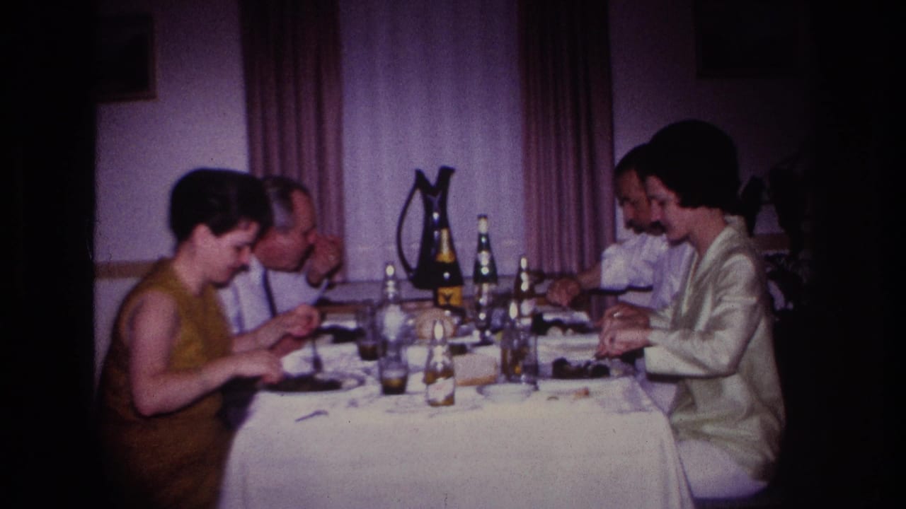 Four adults sit closely around a dining table with plates, bottles, glasses, and curtains in the background.