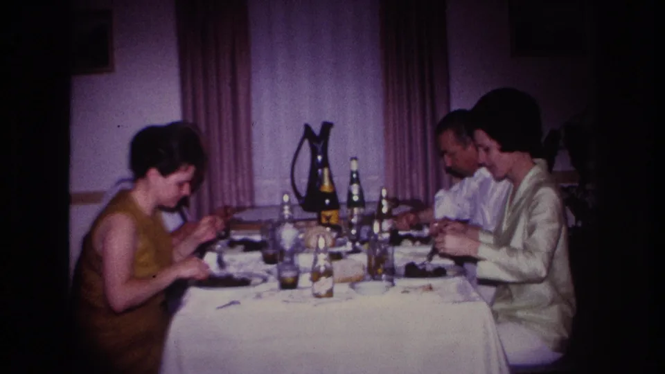 1967 Rockaway Beach home-movie frame showing adults eating at a table in a private interior, with glassware, bottles, plates, and curtains visible.