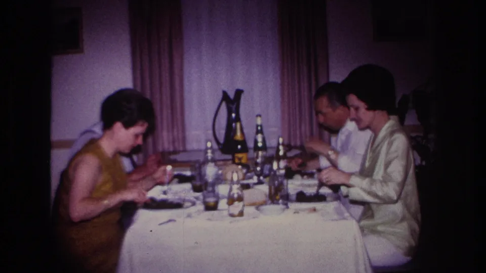 1967 Rockaway Beach home-movie frame of four adults seated at a domestic dinner table, with plates, bottles, glasses, curtains, and soft 8mm color.