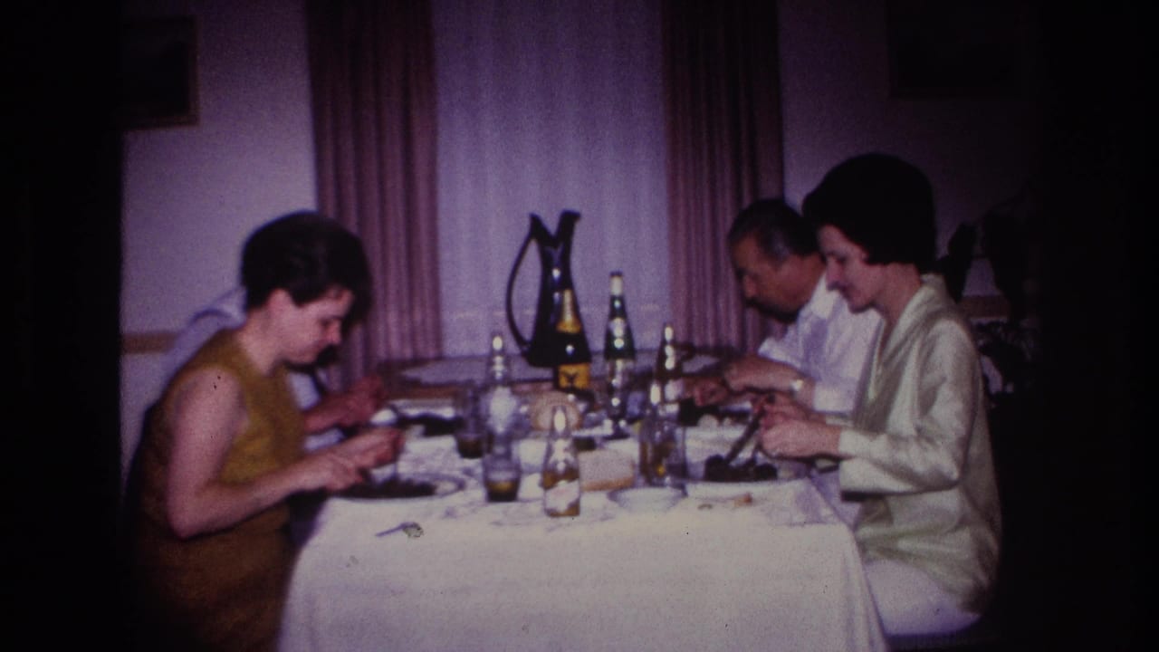 1967 Rockaway Beach home-movie frame showing three adults seated at a dining table with bottles, glasses, plates, curtains, and warm interior light.