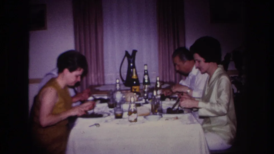 1967 Rockaway Beach home-movie frame showing three adults seated at a dining table with bottles, glasses, plates, curtains, and warm interior light.