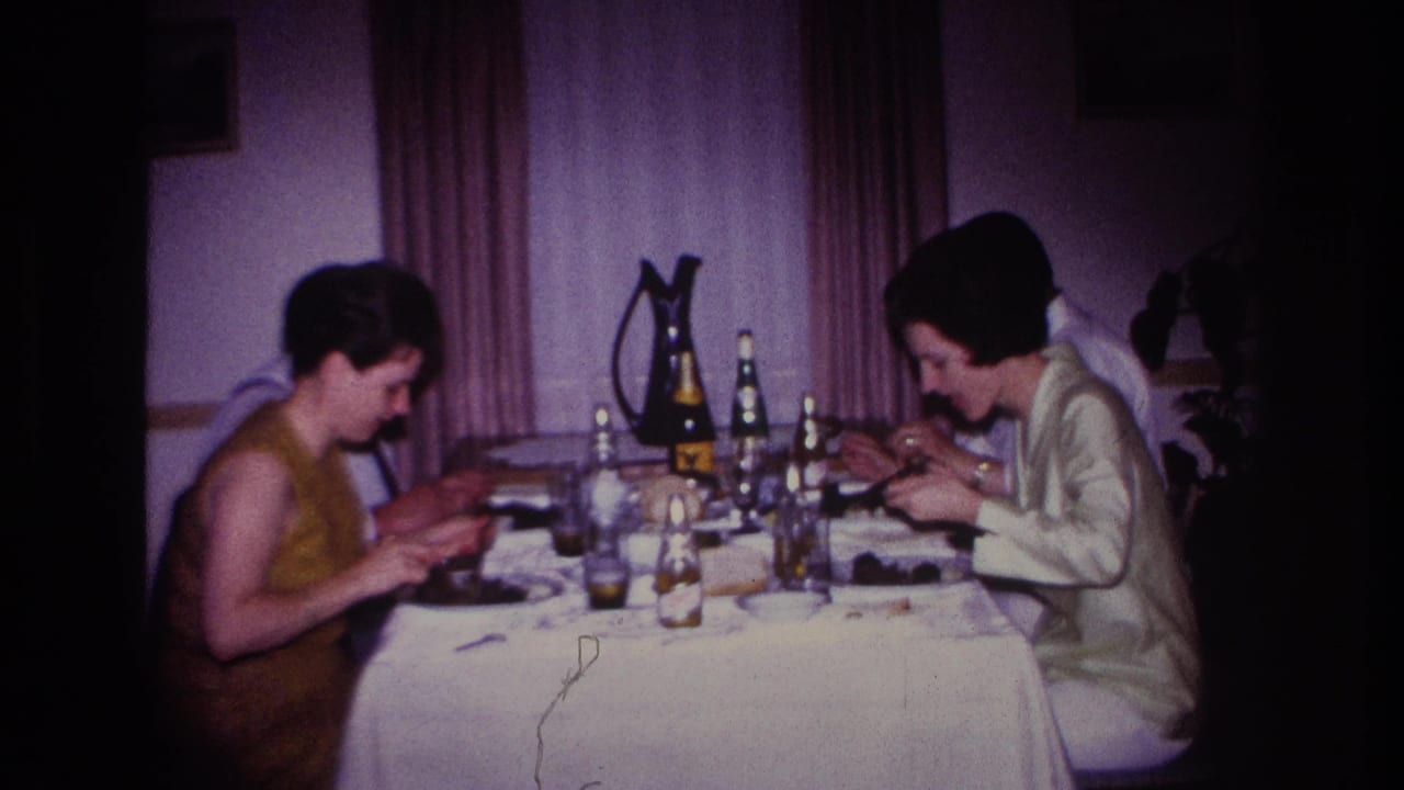 Two women sit at a dinner table with bottles, glasses, plates, and curtains behind them.