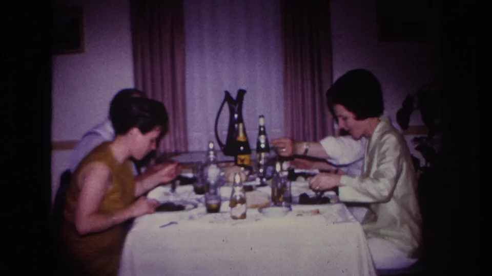 1967 Rockaway Beach home-movie frame of two women at dinner with a partly hidden adult, tableware, bottles, glasses, and curtains in view.