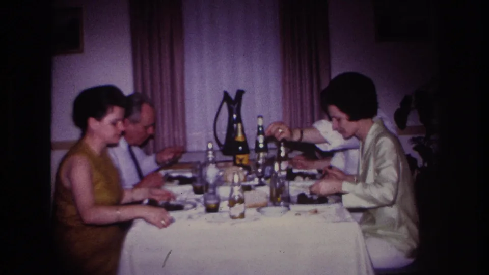 1967 Rockaway Beach dining-room frame showing three adults at a white-covered table with bottles, glasses, plates, and soft curtain backdrop.