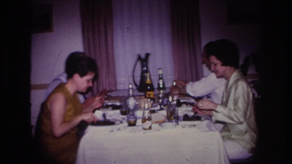 Two women at a 1967 Rockaway Beach dining table appear to eat and drink during a home-movie gathering.