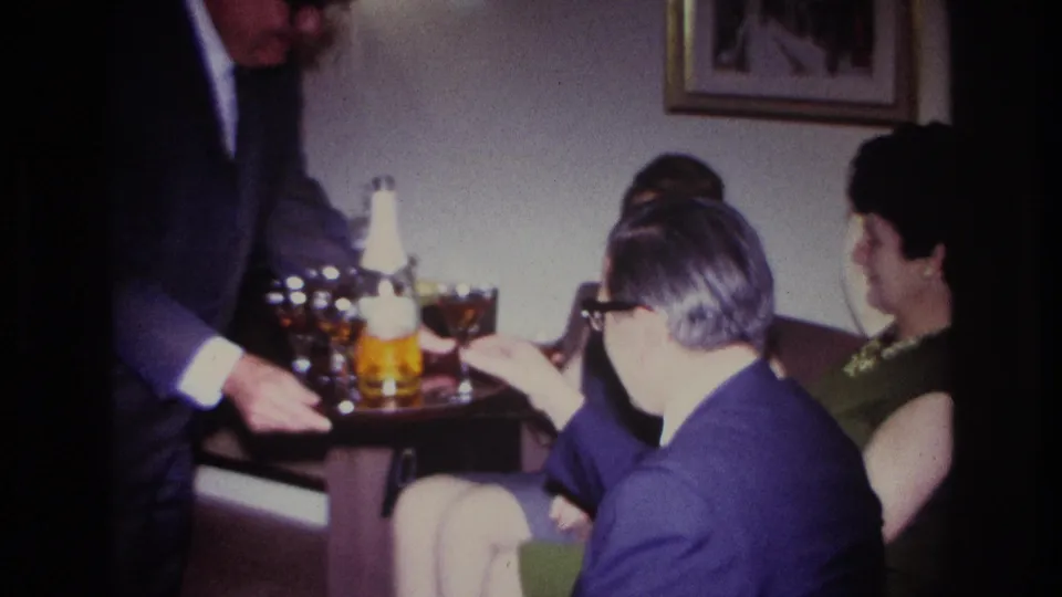 A suited man serves drinks from a tray to seated adults in a 1967 Rockaway Beach home-movie interior.