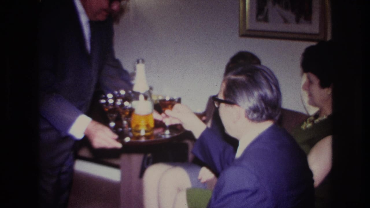 1967 Rockaway Beach home-movie frame showing a suited man offering cocktails to seated adults in a private living room.