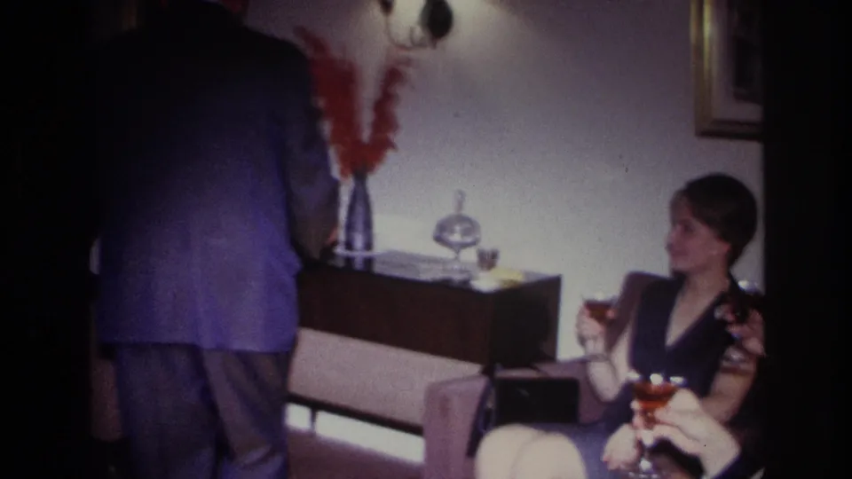 Soft 8mm frame from a 1967 Rockaway Beach gathering, with a standing suited man, seated woman, and cocktail glasses in view.