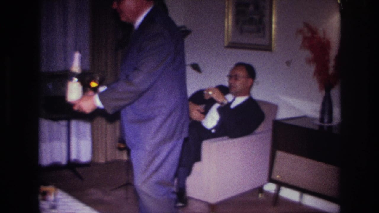 In a 1967 Rockaway Beach home-movie frame, a suited man carries drinks past a seated guest in a living room.