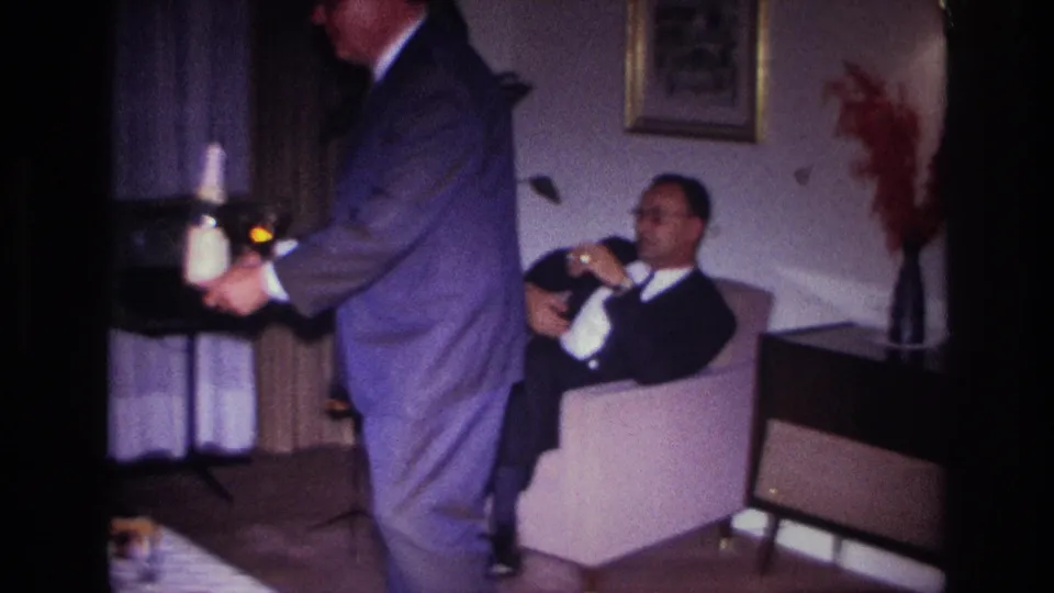 In a 1967 Rockaway Beach home-movie frame, a suited man carries drinks past a seated guest in a living room.