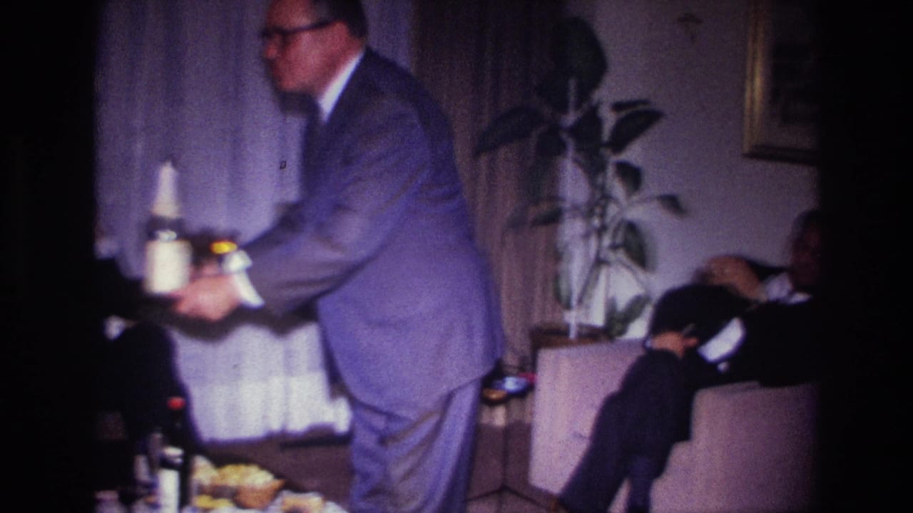 Man in a suit leans forward with a tray near seated guests in a living room.