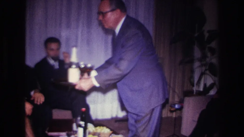 A 1967 Rockaway Beach frame shows a suited man offering glasses from a tray to a seated guest in a private interior.