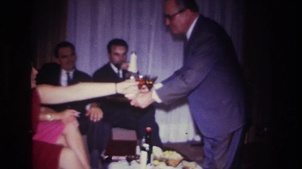 At a 1967 Rockaway Beach living-room party, a woman in a red dress reaches for a glass from a suited man.
