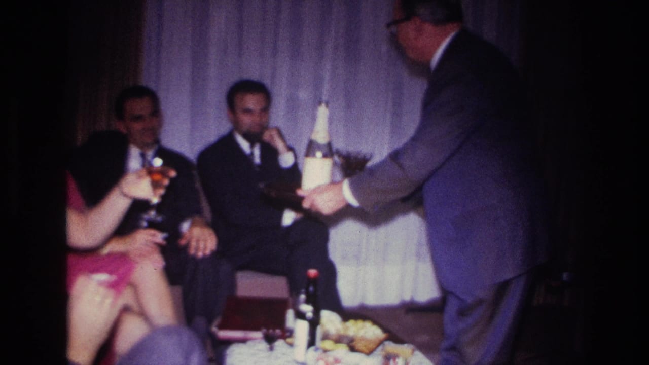 Seated guests hold drinks while a man in glasses presents a tray with cocktails.