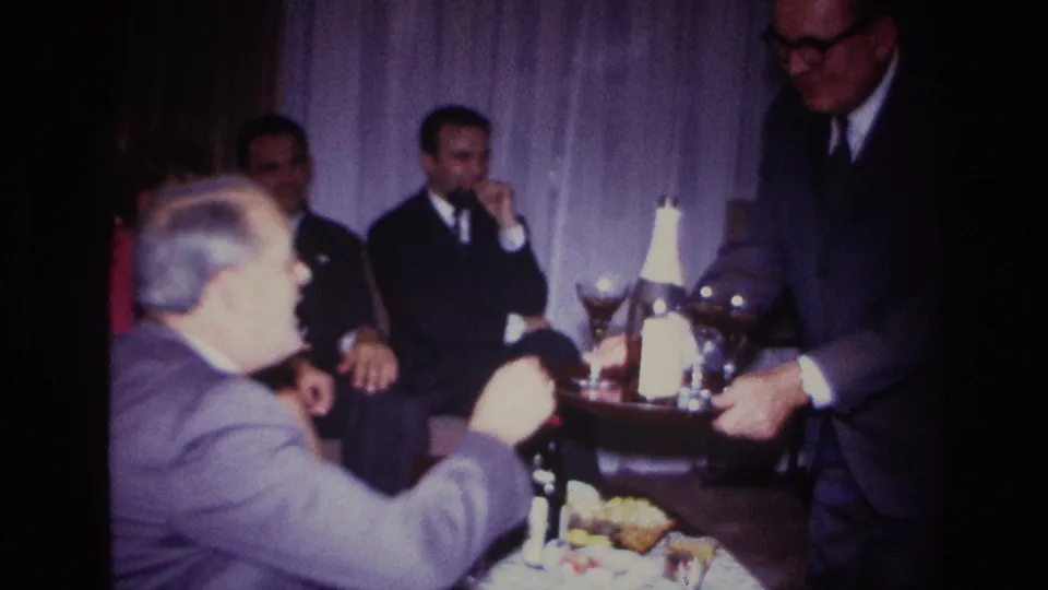1967 Rockaway Beach party frame showing a man reaching toward served cocktails with seated guests behind him.