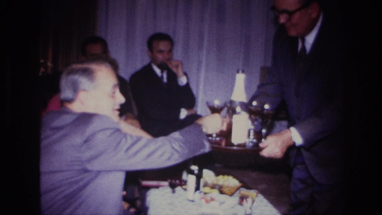 1967 Rockaway Beach home-movie frame showing a server offering drinks to adults seated around a table in a private interior.