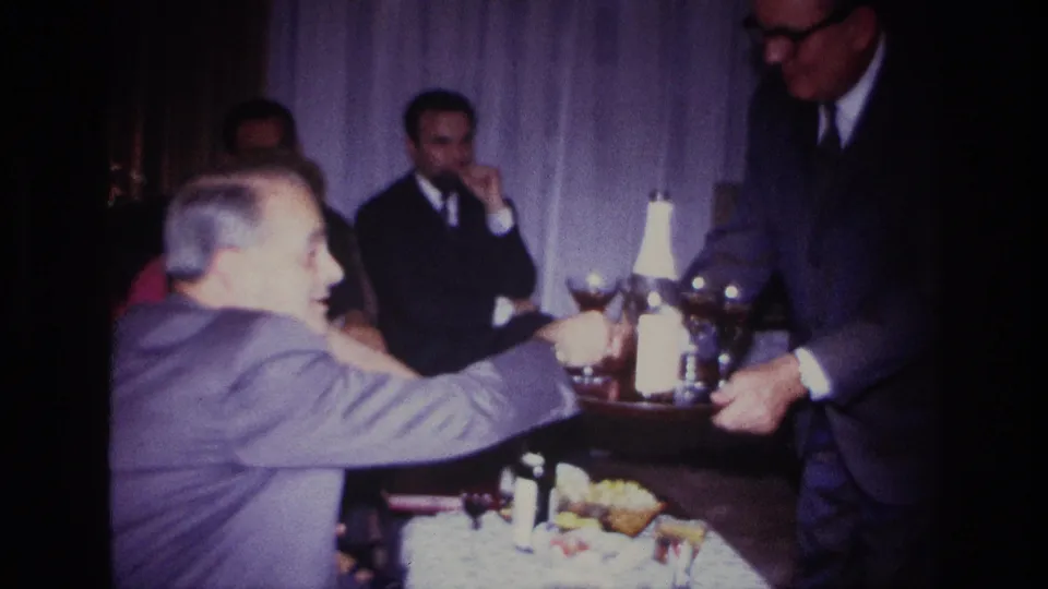 1967 Rockaway Beach home-movie frame showing a server offering drinks to adults seated around a table in a private interior.