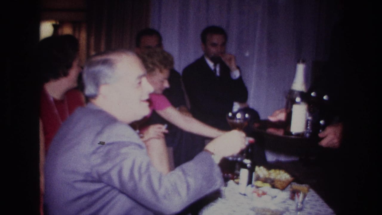 A man in a light gray suit reaches toward a drink while guests sit nearby.