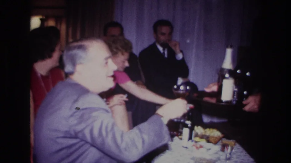 At a 1967 indoor party in Rockaway Beach, a seated man in a light gray suit reaches for a drink as other adults look on.
