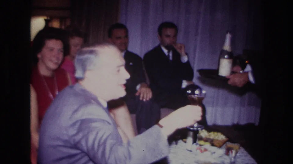 Home-movie frame from the 1967 Rockaway Beach party showing smiling adults at a table while a server offers drinks from a tray.