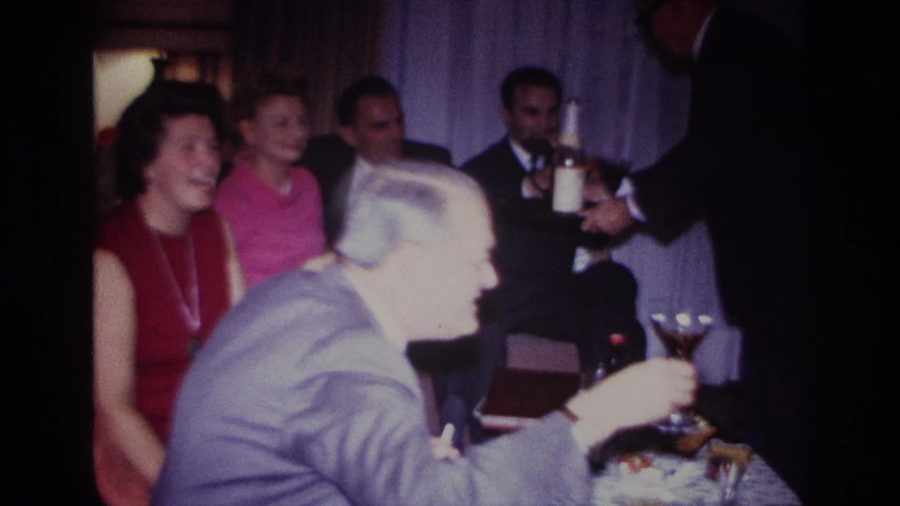 1967 Rockaway Beach home-movie frame showing adults at an indoor gathering, with a man in a gray suit holding a cocktail glass.