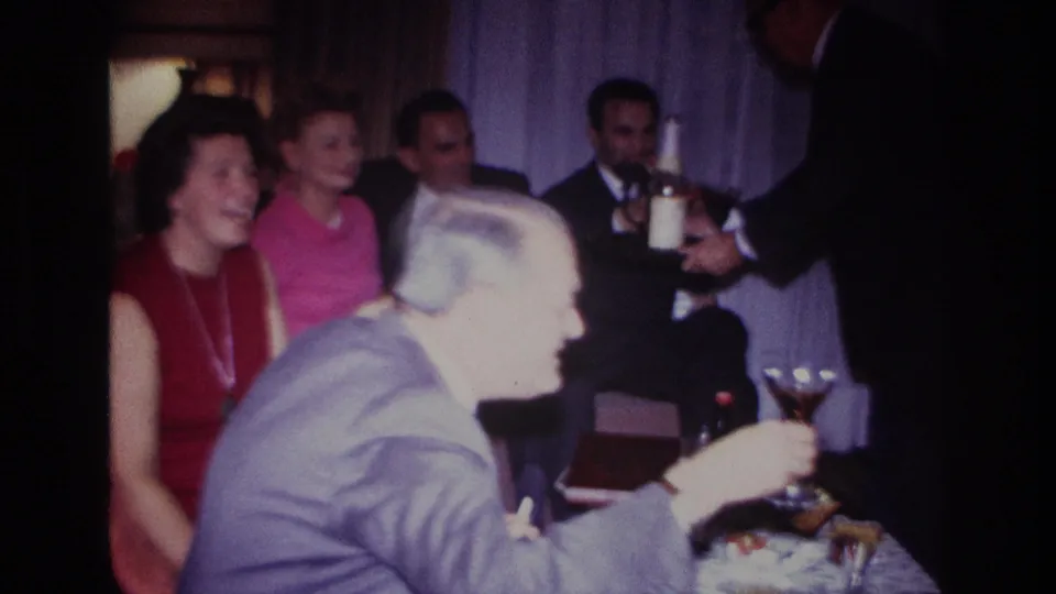 1967 Rockaway Beach home-movie frame showing adults at an indoor gathering, with a man in a gray suit holding a cocktail glass.