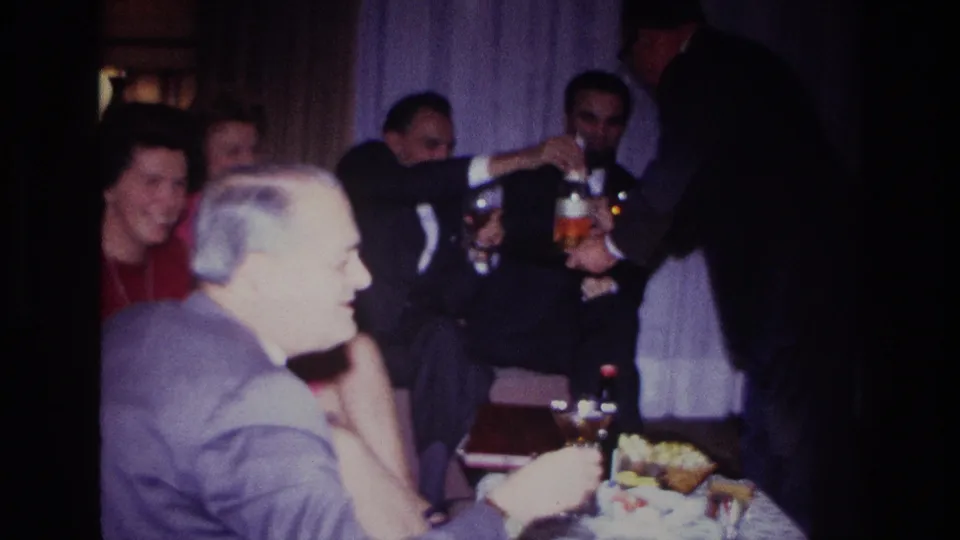 Home-movie frame from the Rockaway Beach gathering showing adults in suits and dresses during a table-side drink pour.