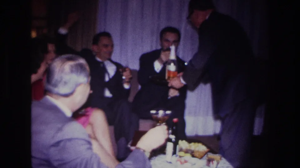 1967 indoor celebration frame with adults in formal clothing, cocktail glasses, snacks, and a server standing at the side.