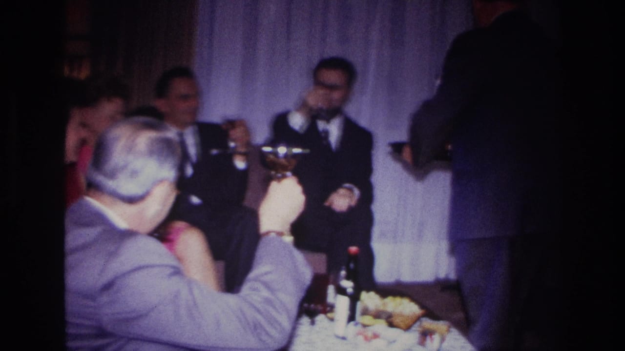 1967 Rockaway Beach home-movie frame of suited men gathered with drinks and food in a curtained private living room.
