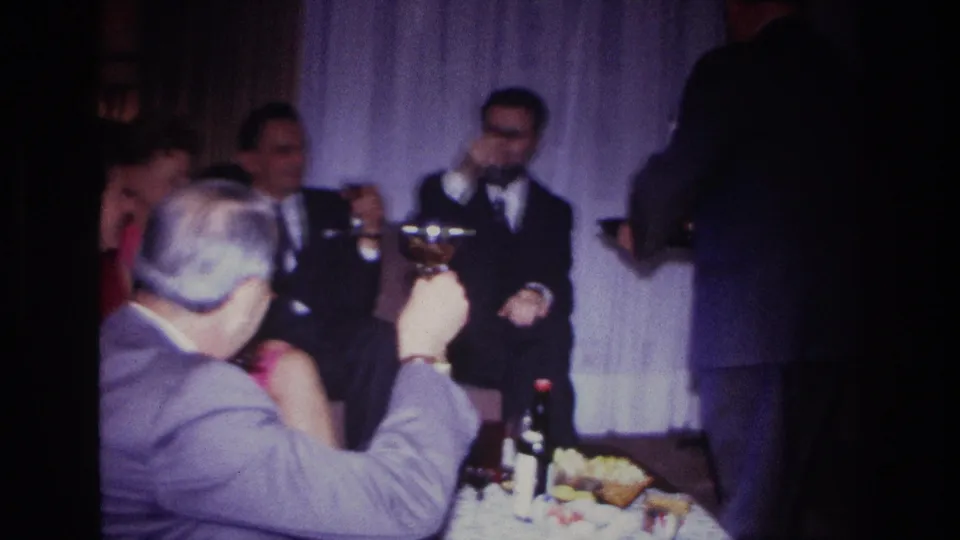 1967 Rockaway Beach home-movie frame of suited men gathered with drinks and food in a curtained private living room.