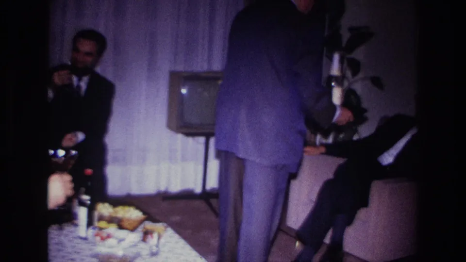 Suited men in a 1967 Rockaway Beach living room hold glasses as a standing figure crosses the handheld frame.