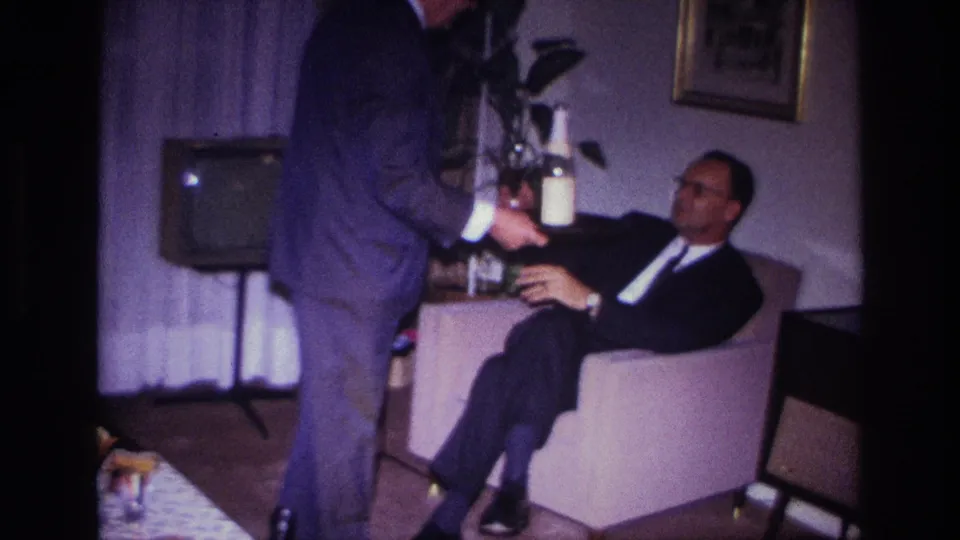 In a Rockaway Beach private interior, a suited man stands with a bottle near a seated guest and mid-century television.