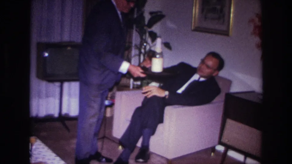 1967 home-movie frame from Rockaway Beach showing a suited guest with a bottle beside a seated man in a private living room.