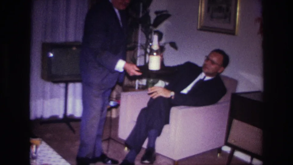 1967 Rockaway Beach home-movie frame showing a suited man serving drinks to a seated man in a private living room.
