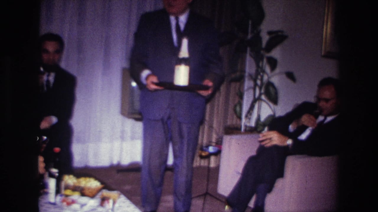 Suited man carries a tray as two other men sit nearby in a living room.