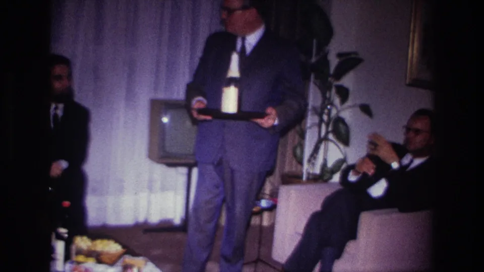 1967 Rockaway Beach home-movie frame showing a suited man with a tray beside a seated guest, food, curtains, and a small television.